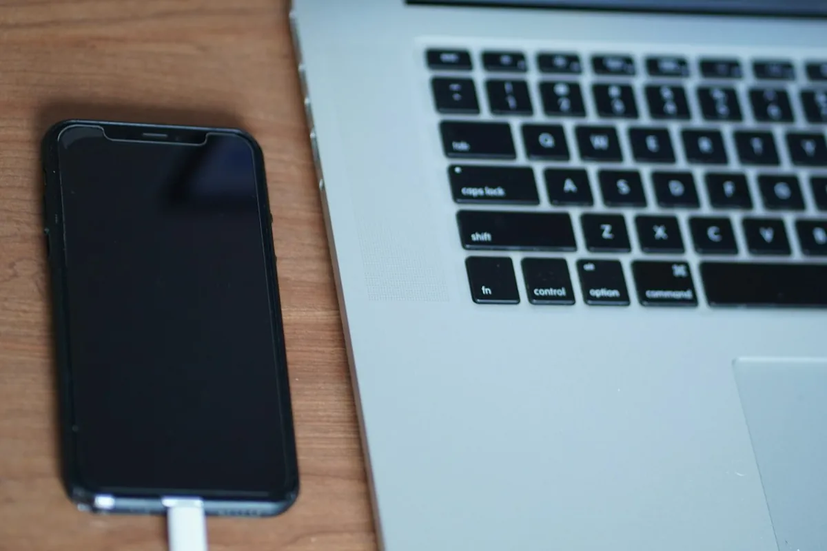 iPhone with a blank black screen on a charging cable beside a laptop, the state a factory reset leaves a phone in for domestic violence digital forensics